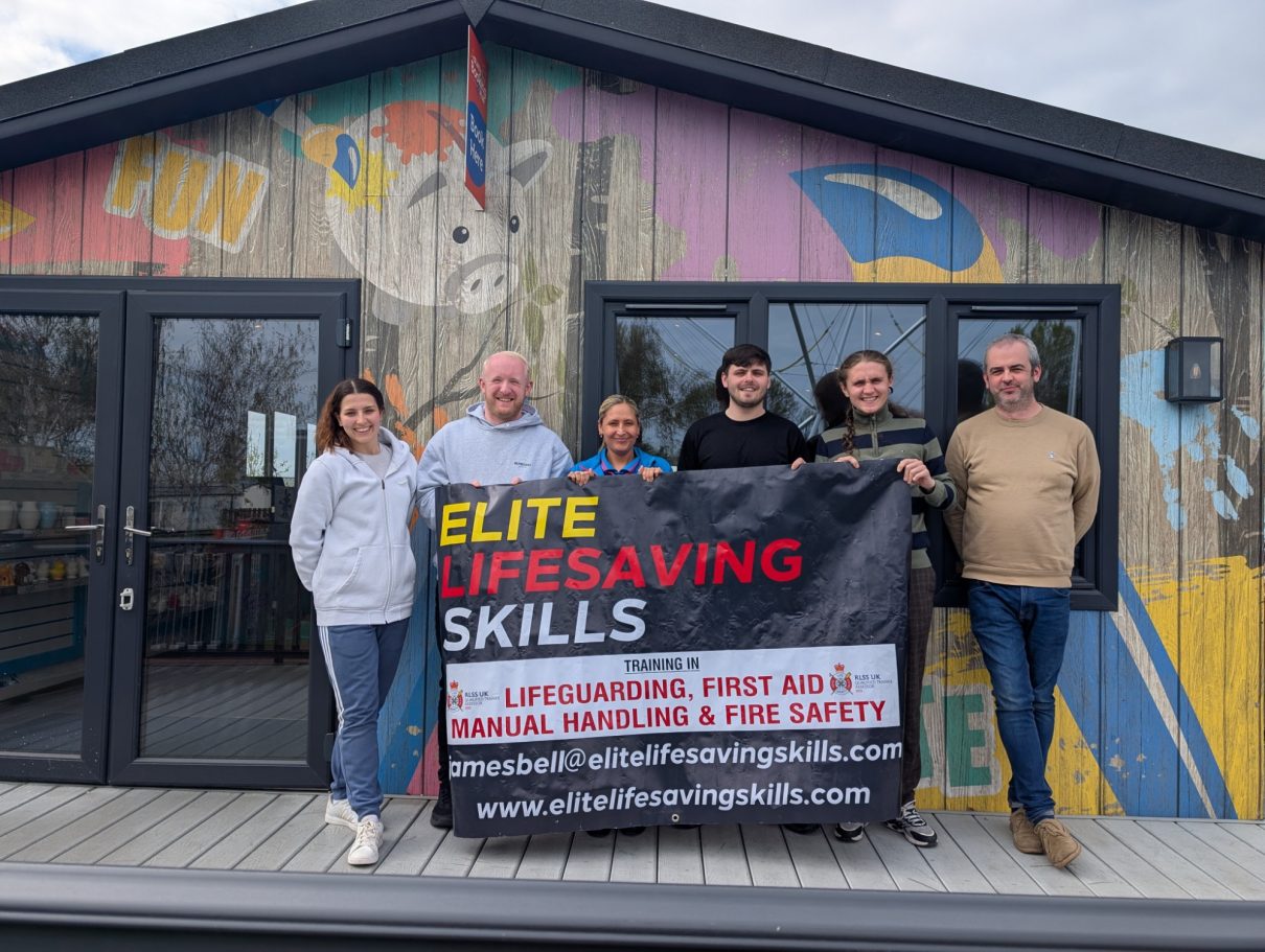 Group of six people holding a banner for Elite Lifesaving Skills in front of a colourful building.